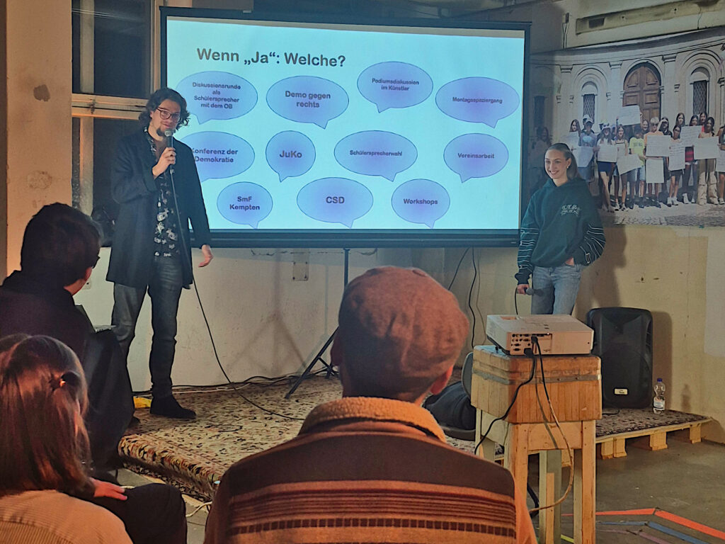 Teilnehmer hören interessiert der Präsentation der Ergebnisse zu. Vorne stehen Silvan Hecht und Anastasia Zoubarev und präsentieren die Antworten, an welchen politischen Veranstaltungen sich kemptener Jugendliche bereits beteiligt haben.