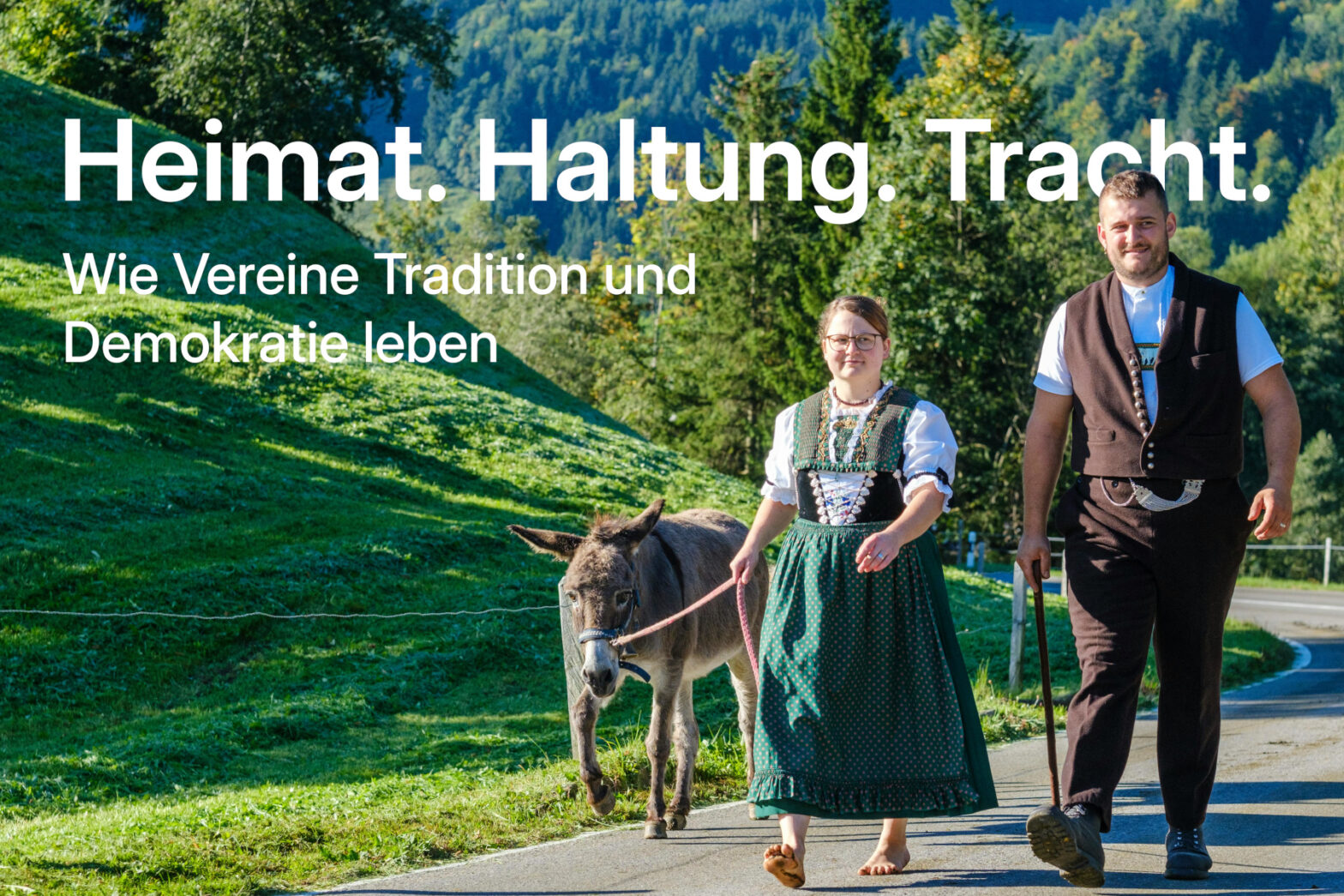 Ankündigungsbild zu Veranstaltung Heimat. Haltung. Tracht. Auf dem Bild laufen ein Dame und ein Herr in Tracht mir einem Esel die Straße entlang