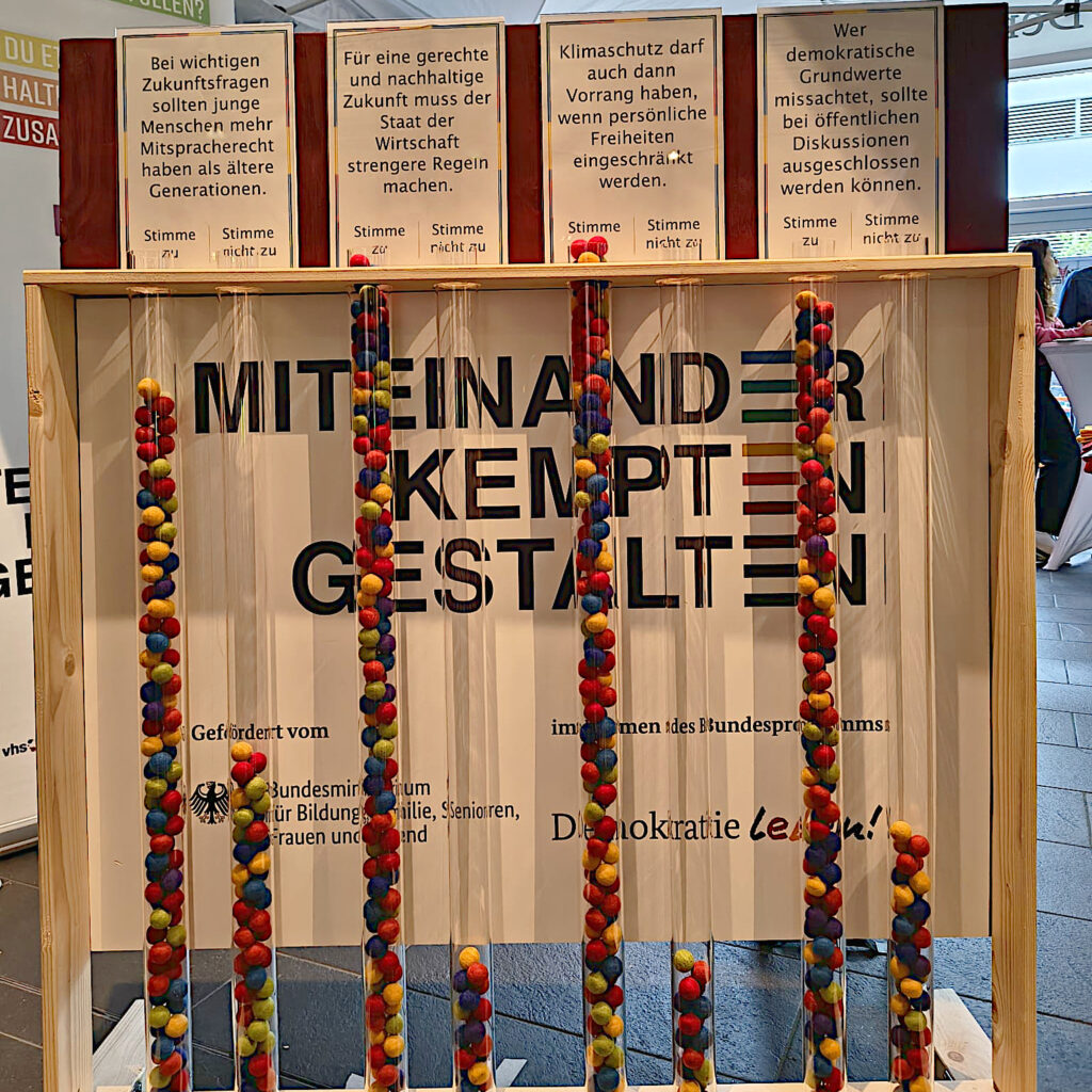 Abstimmungsstation von „Demokratie leben!“ beim Zukunftsfest. Vier Plakate zeigen Aussagen zu Zukunfts- und Demokratiethemen. Darunter stehen durchsichtige Röhren, in denen bunte Filzbälle die abgegebenen Stimmen für Zustimmung oder Ablehnung sichtbar machen.