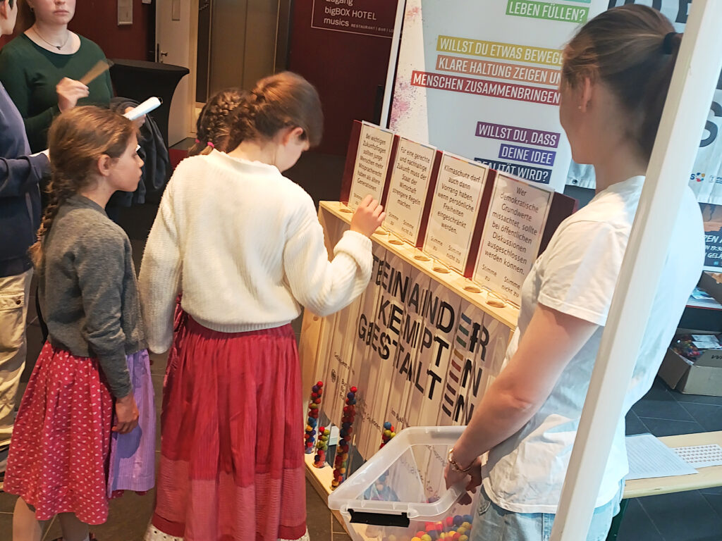 Mehrere Kinder stehen am Mitmachstand von „Demokratie leben!“ beim Zukunftsfest. Sie lesen die Aussagen auf den Abstimmungstafeln und werfen bunte Filzbälle in die Röhren, um ihre Meinung abzugeben.