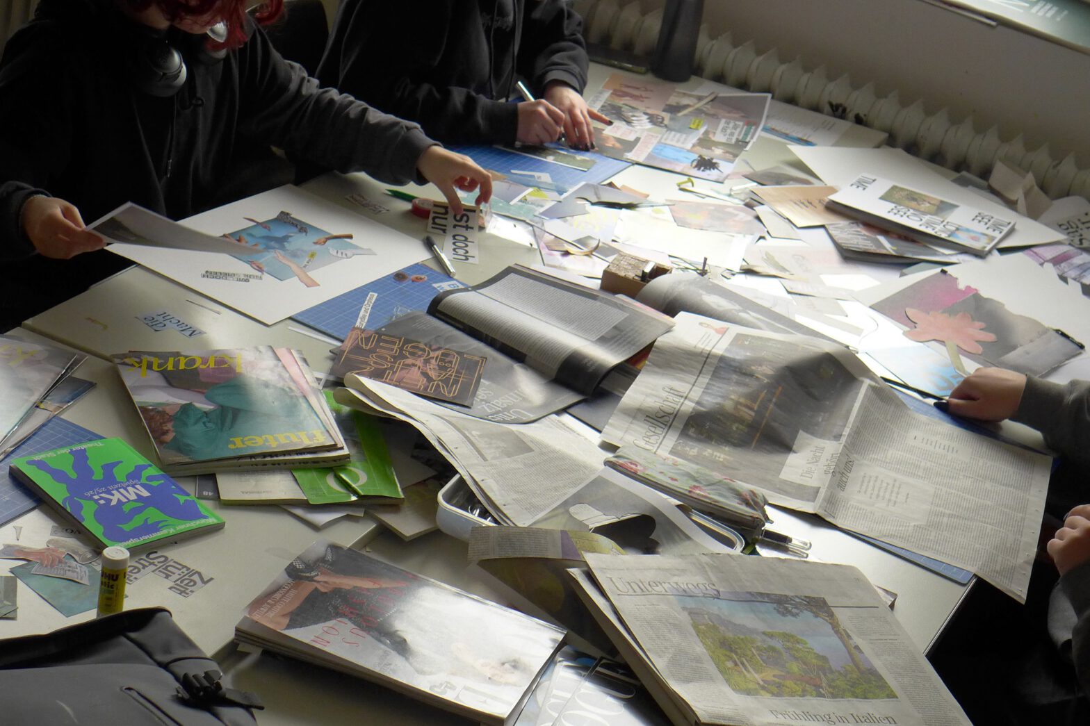 Schülerinnen und Schüler sitzen um Tisch bedeckt mit lauter Zeitungsauschnitten und Zeitschriften. Sie basteln daraus Collagen