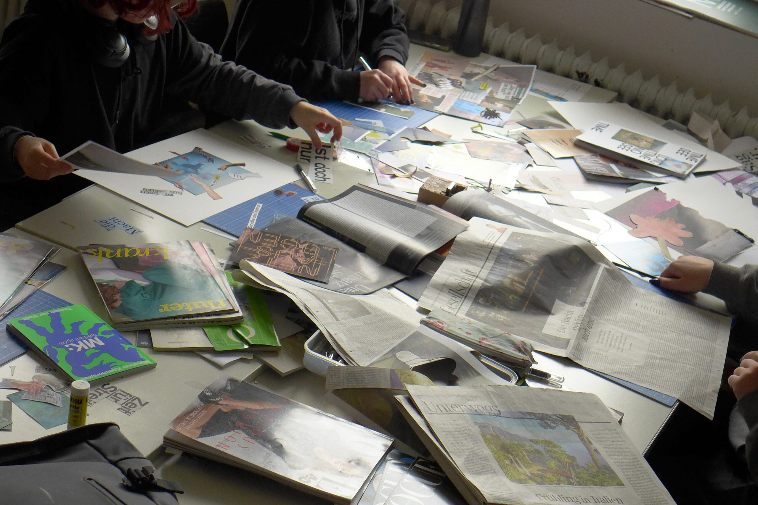 Schülerinnen und Schüler sitzen um Tisch bedeckt mit lauter Zeitungsauschnitten und Zeitschriften. Sie basteln daraus Collagen