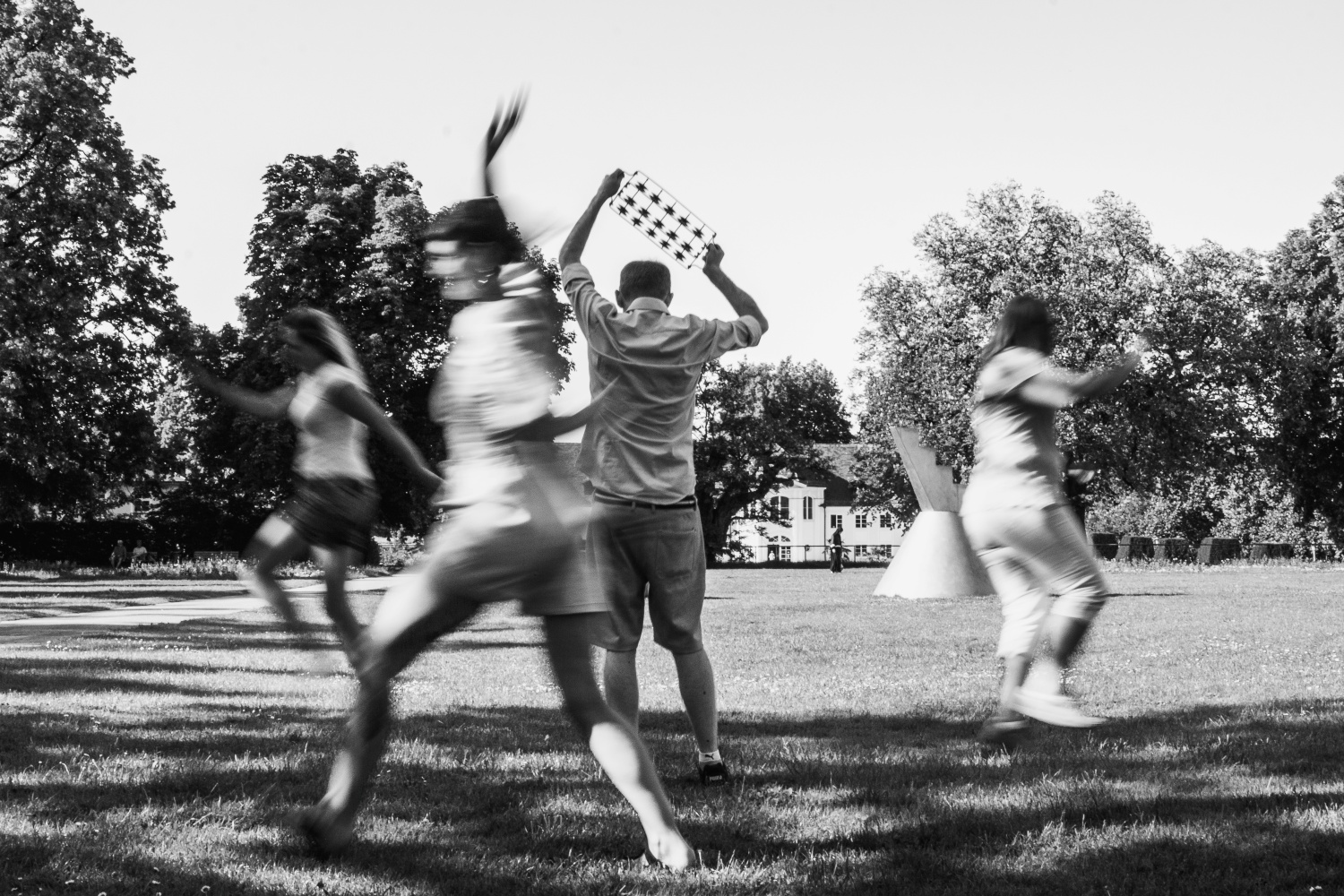 Schwarz-Weiß-Fotografie einer Gruppe junger Menschen, die über eine Wiese laufen. In der Mitte steht eine Person mit dem Rücken zur Kamera und hält eine Rassel über den Kopf; die vorbeilaufenden Personen sind durch Bewegungsunschärfe verschwommen.
