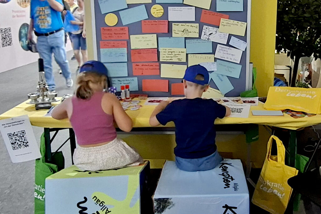 Zwei Kinder gestalten am Mitmachstand eigene Buttons zum Thema Gemeinschaft. Im Hintergrund ist die Wünschewand mit Ideen von Kindern und Jugendlichen für Kempten zu sehen.