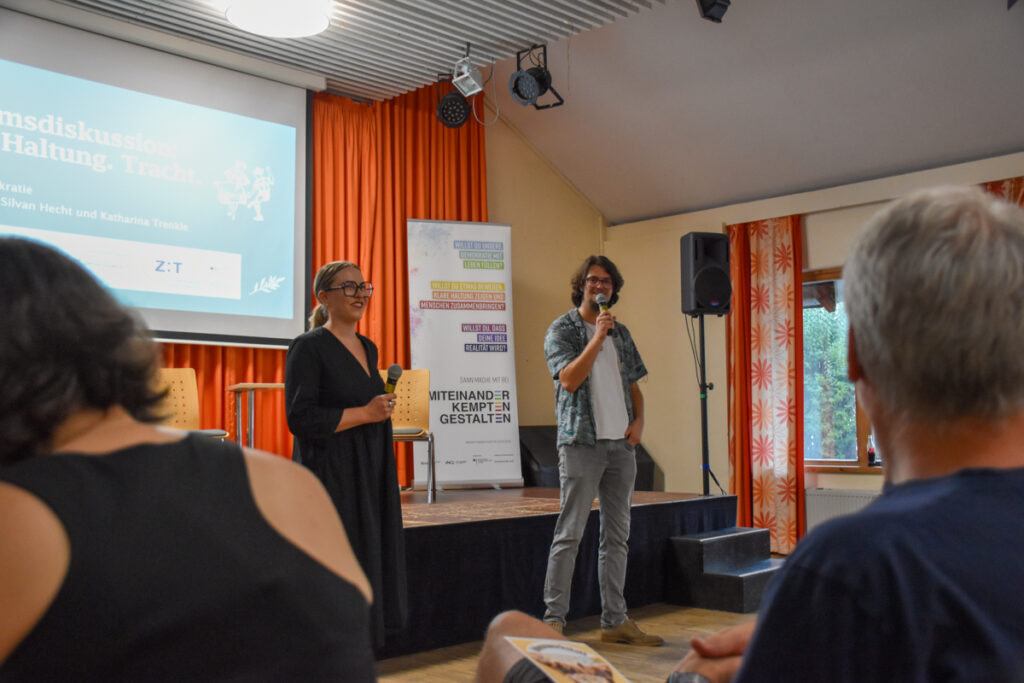 Katharina Trenkle (links) und Silvan Hecht (rechts) begrüßen das Publikum mit Mikrofonen bei der Podiumsdiskussion „Heimat. Haltung. Tracht.“ im Haus International in Kempten.