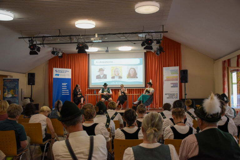 Publikum in Tracht bei der Veranstaltung „Heimat Haltung Tracht“ während einer Podiumsdiskussion mit vier Personen auf der Bühne.