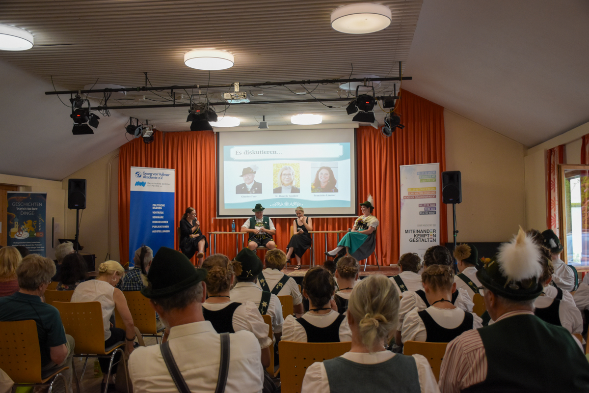 Publikum in Tracht bei der Veranstaltung „Heimat Haltung Tracht“ während einer Podiumsdiskussion mit vier Personen auf der Bühne.