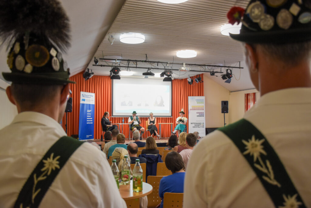 Blick durch zwei Männer in traditioneller Tracht auf die Bühne der Podiumsdiskussion „Heimat. Haltung. Tracht.“ im Haus International in Kempten; davor sitzt das Publikum, auf der Bühne diskutieren die vier Teilnehmenden.