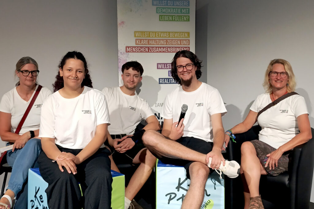Das Team von „Miteinander Kempten gestalten“ sitzt gemeinsam auf der Bühne in Halle 12 der Allgäuer Festwoche und bereitet das Demokratie-Pubquiz vor.