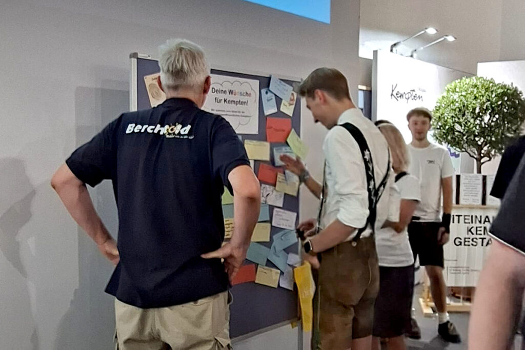 Besucher betrachten die Wünschewand mit Ideen von Kindern und Jugendlichen für Kempten. Im Hintergrund ist der Stand von „Miteinander Kempten gestalten“ zu sehen.